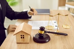 Attorney sitting at desk with scales of justice, gavel and small wooden toy house, working with documents, signing contract agreements. Real estate law, foreclosed property, lawyer services concept