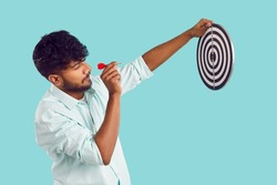 Indian man shooting at target. Profile view of young South Asian man in shirt standing isolated on blue background, holding dart board and aiming little red arrow at bullseye. Setting goal concept