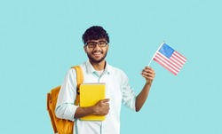 Happy young Indian man studying at American university. Cheerful South Asian exchange program student in glasses with books and bag standing on blue studio background, holding flag of USA and smiling