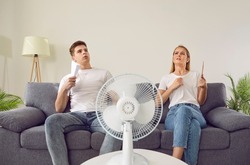 Young couple at home suffering from hot sultry sweltering summertime weather. Sweaty exhausted people waving sheets of paper sitting on sofa with electric fan in overheated house. Summer heat concept