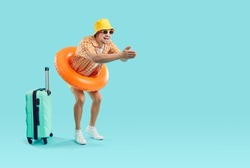 Happy funny young man wearing holiday shirt, summer hat, sun glasses and swim ring standing with suitcase on blue advertising copy space background and getting ready to jump in sea waters