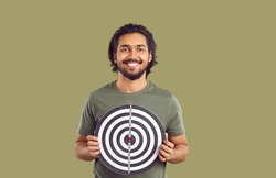 Portrait of happy ethnic man holding shooting target and smiling. Studio shot of young Indian guy in casual T shirt holding dart board with concentric circles and red bullseye. Setting goals concept