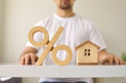 Close up of a wooden percent symbol on a white table with a young man and a little house in the background. Real estate and mortgage interest rates concept