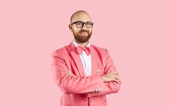 Portrait of smiling young bearded Caucasian male entertainer or performer in suit isolated on pink studio background. Happy man in glasses in party jacket stand with arms crossed.