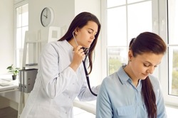 Young female doctor checks woman's breathing and heartbeat at appointment at medical clinic. Focused doctor applies stethoscope to back of female patient in doctor's office. Medicine concept.