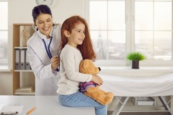 Friendly female pediatrician listens to heartbeat and lungs by applying stethoscope to back of little girl. Cute little girl sitting on table in doctor's office with her favorite toy in her hands.