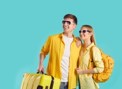 Happy couple with suitcase and travel rucksack go on summer holiday trip. Portrait two joyful people in yellow shirts and sun glasses isolated on turquoise background looking at copyspace on left side