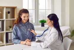 Young woman comes to clinic for heart and lungs checkup. Friendly female doctor sitting at desk in modern medical office, holding stethoscope, listening to patient's breath or heartbeat and smiling