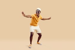 Cheerful stylish african american man having fun dancing on light beige background. Funny smiling dark skinned man in sneakers, shorts, polo shirt and hat with sunglasses. Full length. Banner.