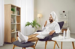 Funny man drinking coffee in morning and enjoying beauty treatments and luxury relaxation at home spa. Man with towel on his head sits on armchair in white bathrobe and with cosmetic mask on his face.