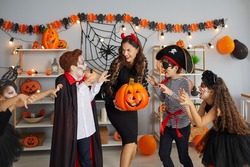 Happy Halloween Day. Funny children jokingly attack young woman with jack-o-lantern in her arms who does not want to give them treats. Kids in creative carnival costumes are scary during Halloween.