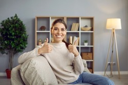 Cheerful satisfied beautiful young woman sitting on sofa in home interior, looking at camera, smiling and doing thumbs up gesture. Happy house owner or online course participant shows her satisfaction