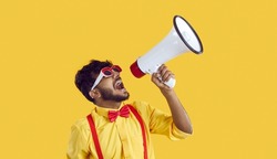 Screaming in megaphone. Funny Indian man in humorous clothes makes loud advertisement on vivid yellow background. Guy in yellow shirt, red suspenders and bow tie party glasses announces information
