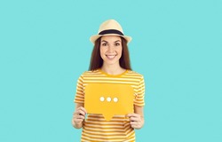 Positive cheerful young woman holding speech bubble sign with three dots isolated on light blue background. Happy caucasian woman in t-shirt and summer hat holding social media sign looking at camera.