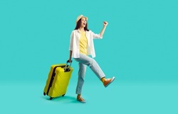Happy woman with yellow suitcase going on summer holiday. Smiling overjoyed young girl wearing jeans, shirt and panama hat walking with her travel bag on bright turquoise background. Vacation concept