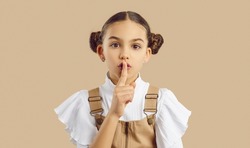 Little kid doing silencing gesture. Child hiding important secret. Beautiful girl with cute hair buns looking at camera, holding her finger on lips and asking you to not share top secret information