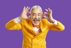 Portrait of funny mature man in eccentric suit isolated on purple studio background show casino chips. Smiling old male croupier or poker player enjoy gambling. Hobby and entertainment.