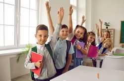 Happy clever school children ready for class. Small group of cheerful cute smart kids in casual clothes with books and backpacks standing near row of desks in classroom, raising up hands and smiling
