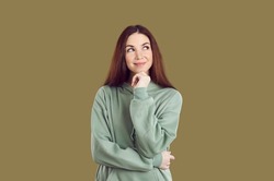 Sly young woman with smile ponders over idea, looking aside on khaki background. Joyful caucasian woman in casual hoodie touching her chin and looking at upper right corner with sly expression.