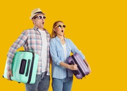 Surprised young Caucasian man and woman tourists in sunglasses holding suitcases for travel dumbfounded by lucrative invitation to tour or cheap offer from airline company stand on yellow background