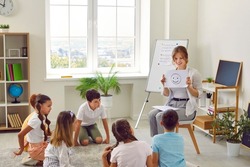 We learn to talk about emotions. Friendly female school psychologist talks about emotions during meeting with group of elementary school students. Woman shows smiling emoticon to students.