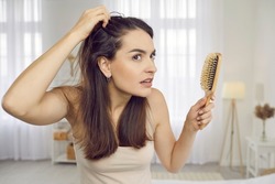 Portrait of woman in her 20s or 30s looking at her reflection with scared nervous expression as she notices bad signs like scalp dandruff, hair thinning, or hair falling out. Hair loss problem