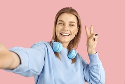 Happy cheerful confident young online social media blogger girl takes selfie photo on pink color background. Positive high school student holding mobile phone, smiling and doing peace victory