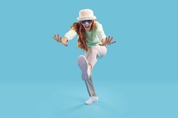 Cheerful crazy and strange young woman having fun on light blue background in studio. Excited Caucasian young woman in stylish casual clothes and sunglasses joyfully shouting while dancing. Web