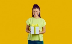 Smiling young Caucasian woman hold wrapped gift congratulate with birthday anniversary. Happy girl isolated on yellow studio background with giftbox greet make surprise. Celebration concept.