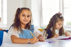 Portrait of cute smiling little schoolgirl sitting in classroom and enjoying her studies. Smiling little girl with pencil and notebook is sitting at desk in classroom and looking at camera.