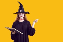 Serious creepy woman in witch character pointing with finger to side on orange background in studio. Portrait of woman with bloody makeup in black dress and witch's hat holding spell book. Web