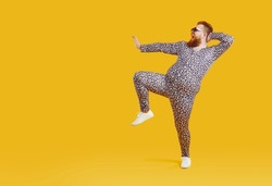 Funny fat fashion guy in pajamas posing on copy space background. Full length portrait of happy joyful plump bearded young man wearing leopard PJs dancing on blank yellow copy space background