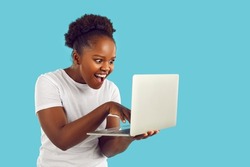 Funny dark-skinned woman with crazy facial expression looks at laptop screen on light blue background. Cheerful emotionally affected woman typing on laptop looking for crazy discounts.