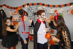 Halloween children's makeup. Young woman prepares children for costume party and applies makeup to little boy making him vampire. Boys and girls have fun in room with Halloween decor.
