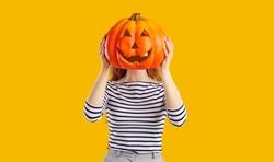 Studio portrait of a woman with a pumpkin head. Funny young girl standing isolated on a yellow background, holding a carved orange pumpkin and hiding her face behind it. Halloween concept