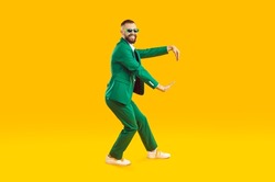 Funny young guy dancing in a cool party outfit. Happy, joyful man wearing a stylish green suit and sunglasses dancing isolated on a bright yellow colour background. Full body studio shot, side view