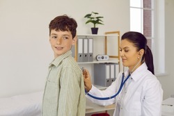 Child getting a professional health exam at the doctor's office. Young woman who works as a pediatrician at the clinic holding a stethoscope and checking lungs or heartbeat of a little school boy