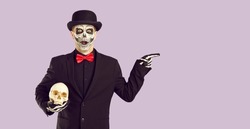 Man in Halloween costume with surprised, scared and shocked expression is pointing at advertising space. Man in suit and Halloween make-up holds skull and points to copy space on lilac background.