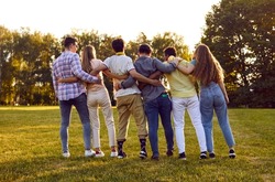 Group of happy friends meet on a nice summer day. Backside shot of several young men and women huddling in park. Back view of diverse people standing in row on green grass lawn and hugging each other