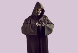 Man dressed in scary Halloween costume. Studio portrait of Mr Death with skeleton skull makeup on face wearing black cape with hood looking at camera while standing isolated on light purple background