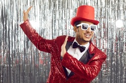 Portrait of a happy funny young black man in a red sequin party jacket, top hat and sunglasses having fun and dancing against a silver foil fringe background