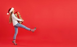 Full length profile side view happy excited child in cap and cute striped socks takes wide step while carrying stack of Christmas gift boxes on red copyspace banner background. Buying presents concept
