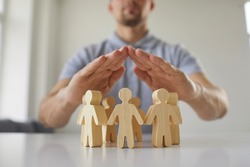 Young man holding hands above small wooden toy human figures placed on white desk as metaphor for human rights protection and safe community of people. Close up, closeup. Society, care, safety concept
