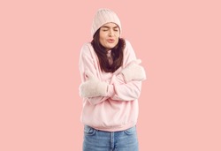 Woman freezing on very cold winter day. Young brunette lady in warm light pink hat, sweatshirt and mittens standing on pastel pink background, feeling cold and shivering with funny face expression