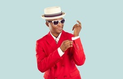 Young trendy African American man in red suit and hat for attending party or fashionable popular event, adjusts sleeves and prepares for work of showman or presenter standing in pastel blue studio