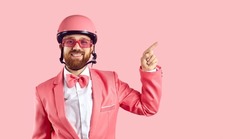 Portrait of funny man with ginger beard and happy face expression wearing pink bike helmet, suit, bowtie and sunglasses standing isolated on pink background, smiling and pointing to side at copy space