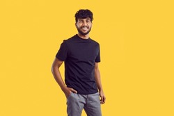 Young cheerful attractive South Asian man with dark curly hair posing with smile in casual clothes holding hand in pocket of trousers stands on solid yellow background in studio. Ethnic Indian human