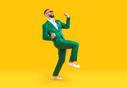 Young man celebrating success. Happy funny joyful excited guy in stylish green party suit and cool glasses raising fist up and dancing isolated on bright yellow background. Full length shot, side view