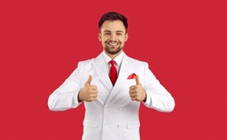 Happy attractive young man in elegant white suit standing isolated on solid red background, smiling and showing thumbs up. Handsome salesman guaranteeing good service and best quality products