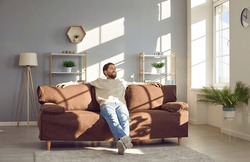 Man relaxing on the sofa at home. Happy young man sitting on a comfortable brown couch in a spacious living room interior with modern furniture, house plants and big windows
